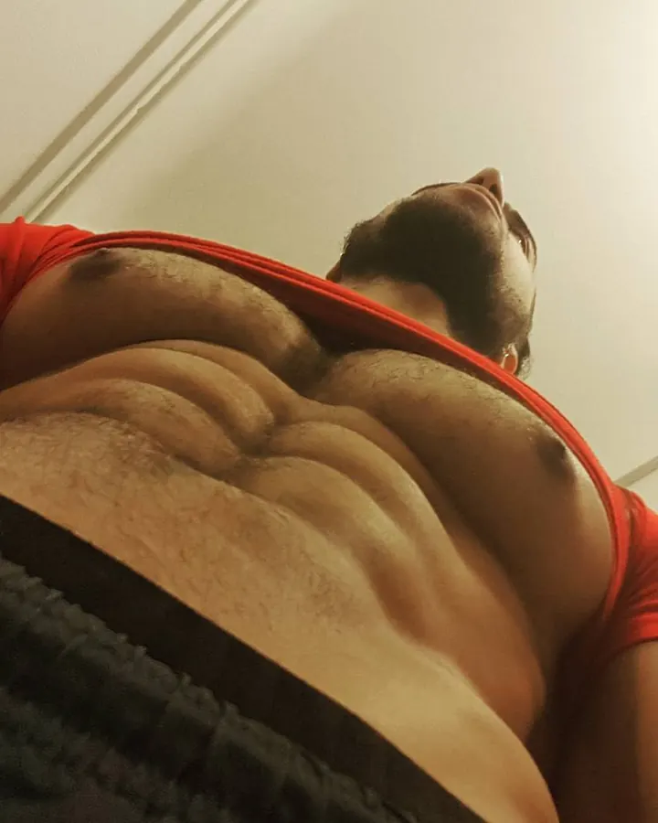 Homme barbu en t-shirt rouge, allongé, regardant vers le haut, torse musclé visible, peau foncée.