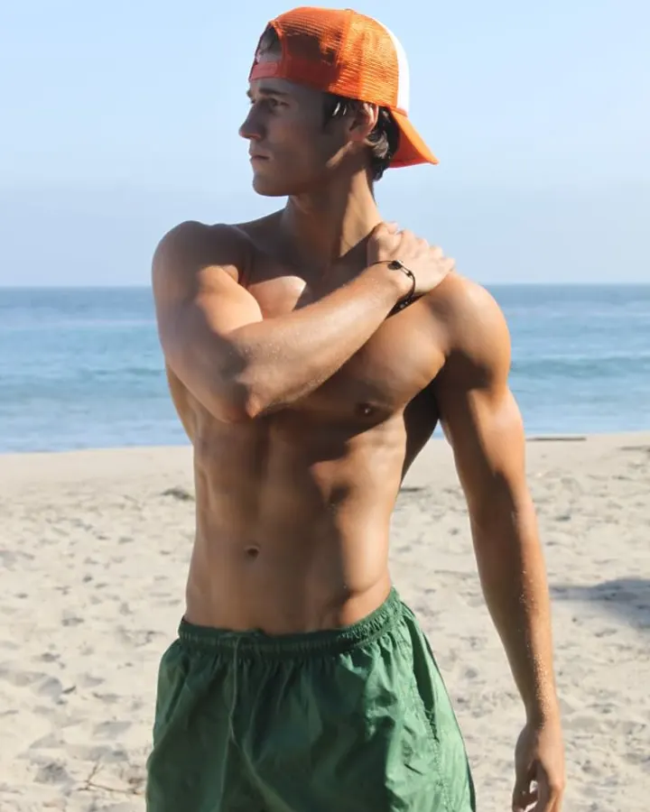 Homme torse nu sur une plage, portant un short vert et une casquette orange, regardant vers l'horizon