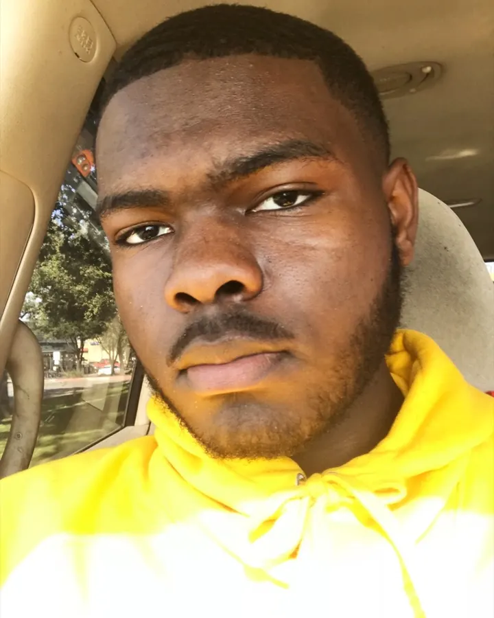Homme noir en hoodie jaune, prenant un selfie dans une voiture, regardant directement la caméra, avec des arbres en arrière-plan.
