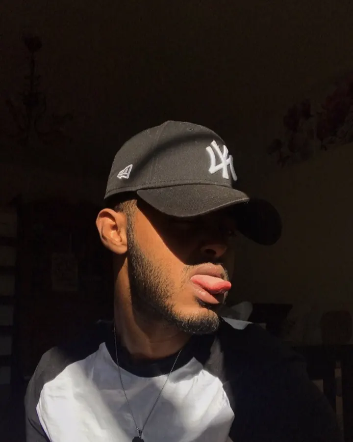 Homme noir en chemise noire, portant des lunettes de soleil, une couronne verte, un haut rayé noir et blanc, un pantalon rayé noir et blanc, les mains sur le torse, debout sur une colline herbeuse.