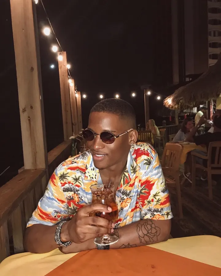 Homme noir souriant, portant des lunettes de soleil rondes, une chemise hawaïenne colorée, tenant un verre de boisson, assis à une table en extérieur la nuit, avec des guirlandes lumineuses en arrière-plan.