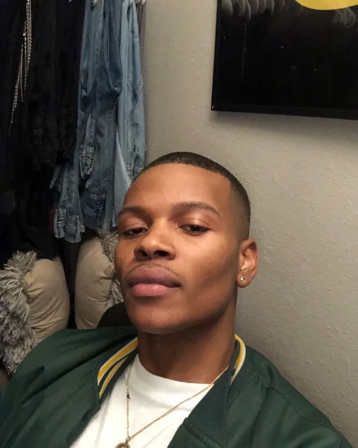 Homme noir en veste verte à col jaune, portant un t-shirt blanc, un pendentif, un piercing à l'oreille, regardant directement la caméra, devant un mur gris avec des vêtements suspendus.