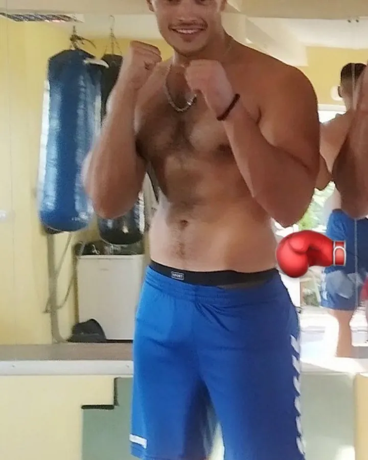 Homme torse nu, en short de boxe bleu, poings serrés, souriant, dans une salle de sport.
