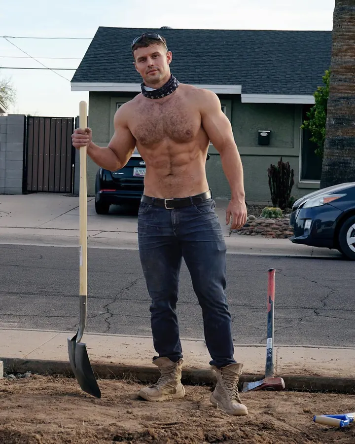 Homme torse nu, tenant une pelle, en jeans et bottes, devant une maison.