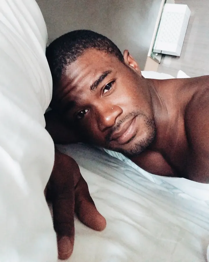 Homme torse nu, allongé sur un lit, regardant l’appareil photo, main posée sur le drap.