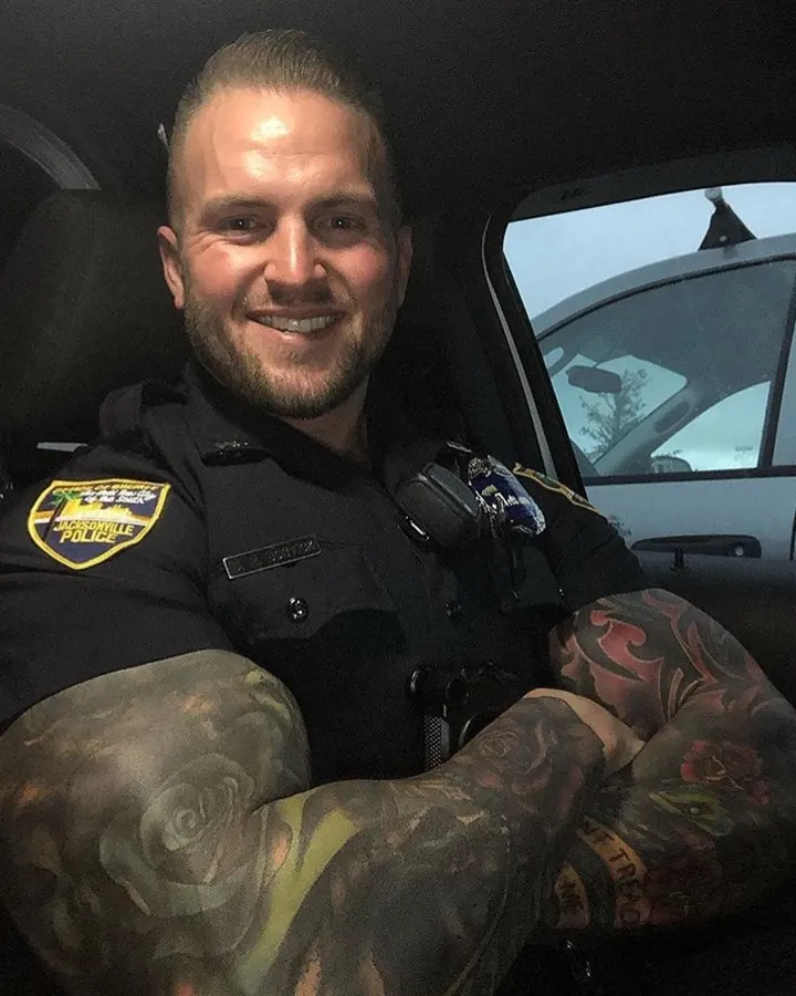 Homme en uniforme de police, souriant, bras croisés, tatouages visibles.