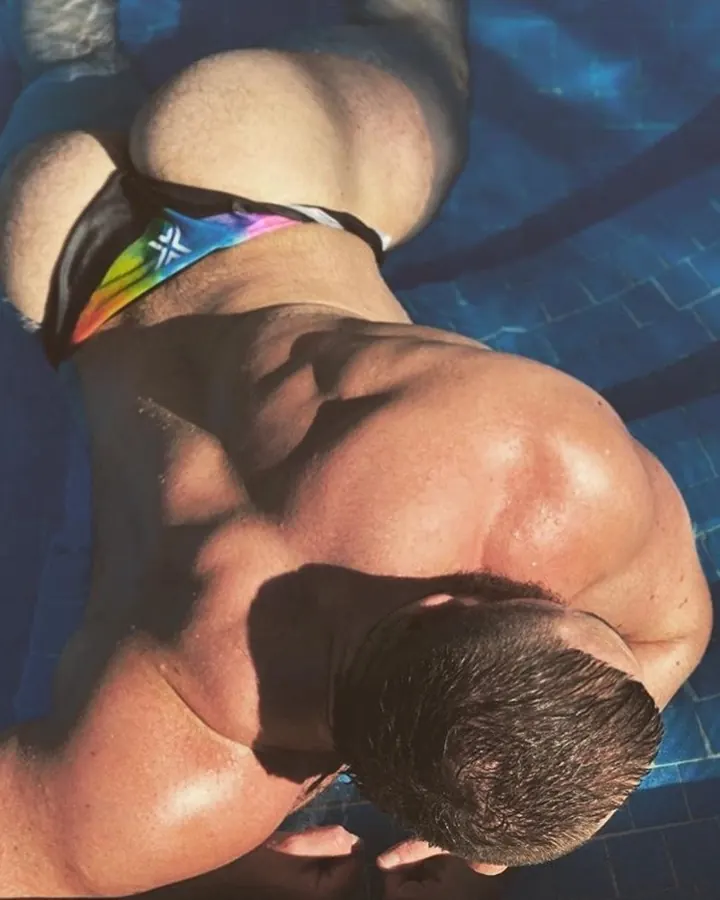 Homme européen barbu, torse nu, portant un slip bleu, posant sur une terrasse ensoleillée.
