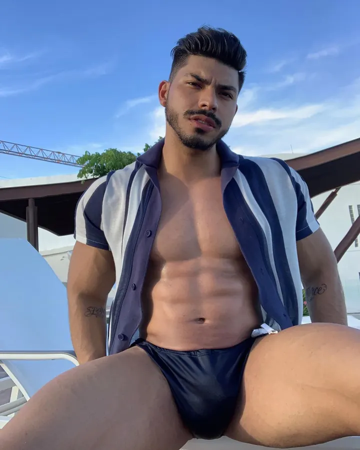 Homme latino barbu, torse nu, portant un slip noir, posant dans une piscine.