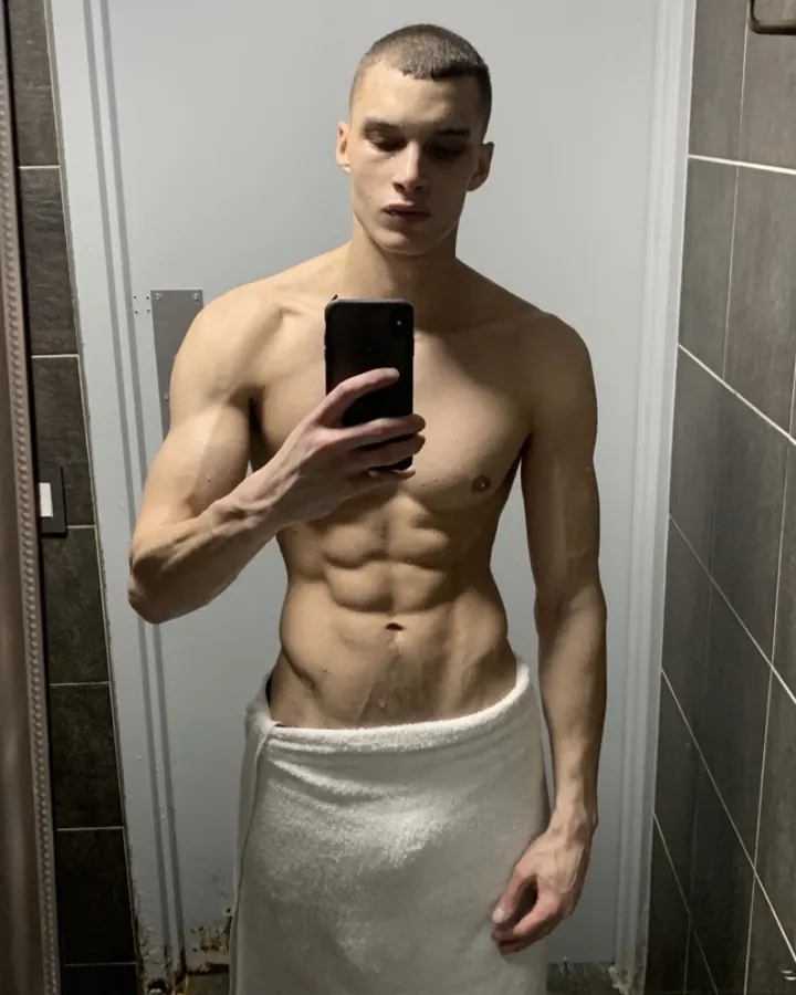 Jeune homme européen torse nu, prenant un selfie dans une salle de bain, enveloppé dans une serviette.