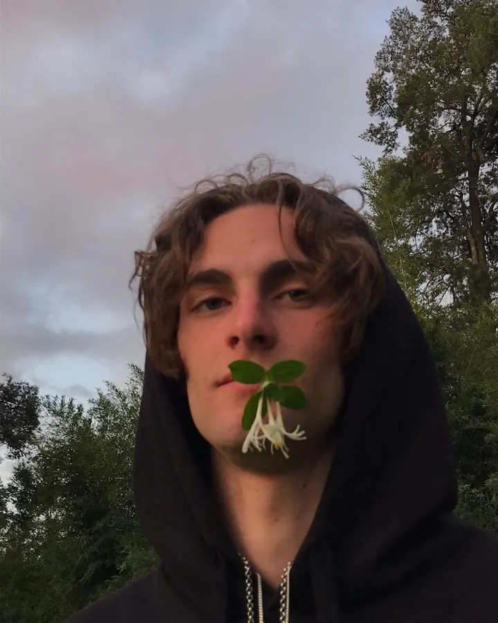 Jeune homme aux cheveux bruns, portant un sweat à capuche noir, tenant une fleur blanche entre ses lèvres