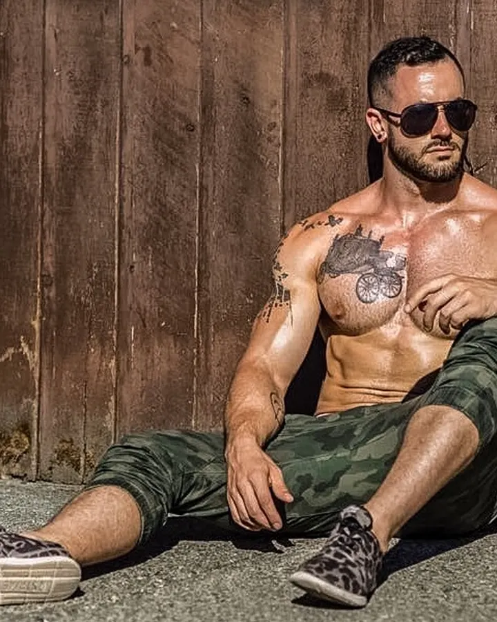 Homme torse nu, assis contre un mur en bois, portant des lunettes de soleil et des pantalons camouflage, tenant une cigarette
