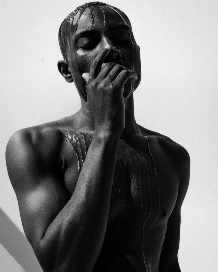 Homme noir, torse nu, couvert de liquide brillant, main sur le visage.