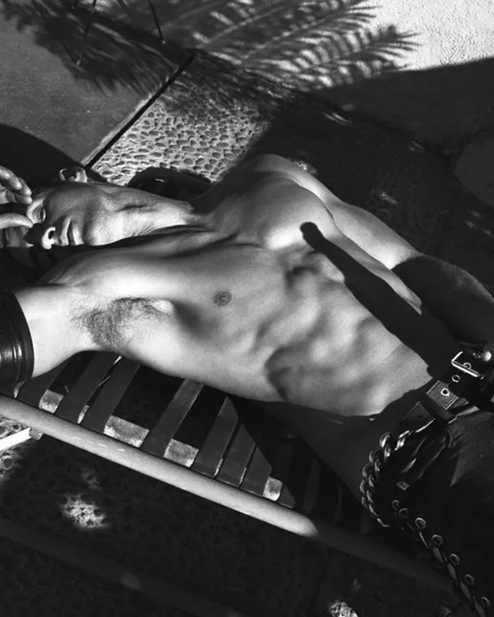 Homme torse nu, aux cheveux bruns, posant devant un sac de frappe, regard intense.