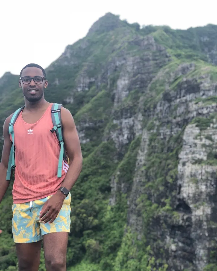 Homme en tenue de randonnée, souriant, debout devant une montagne verdoyante, sac à dos vert.