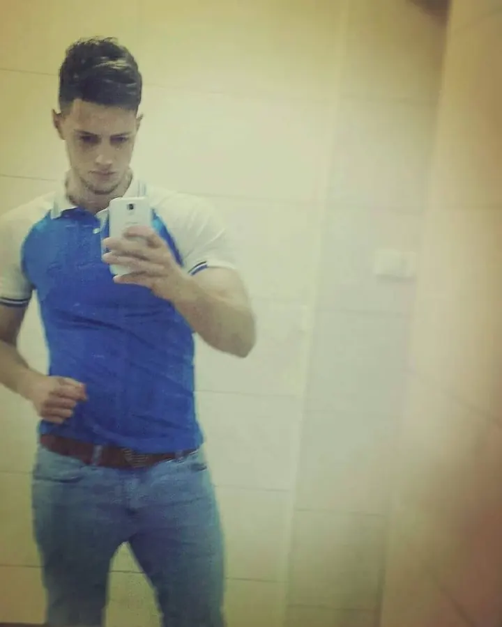 Jeune homme en polo bleu et blanc, prenant un selfie devant un mur beige, cheveux coiffés, regard concentré.
