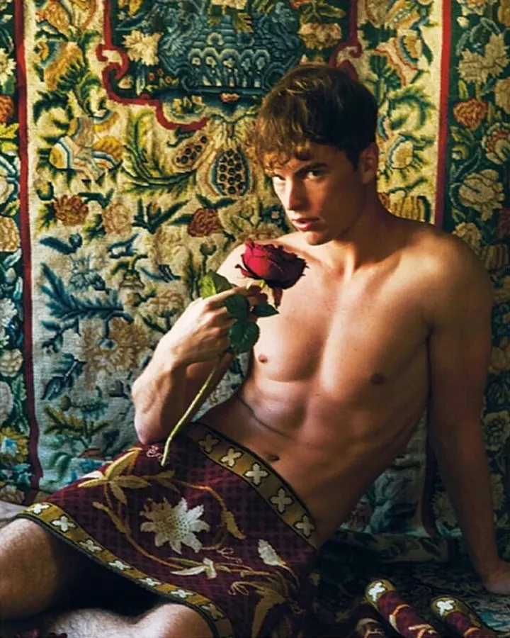 Homme torse nu, assis sur un tapis décoratif, tenant une rose rouge, regardant l'appareil.