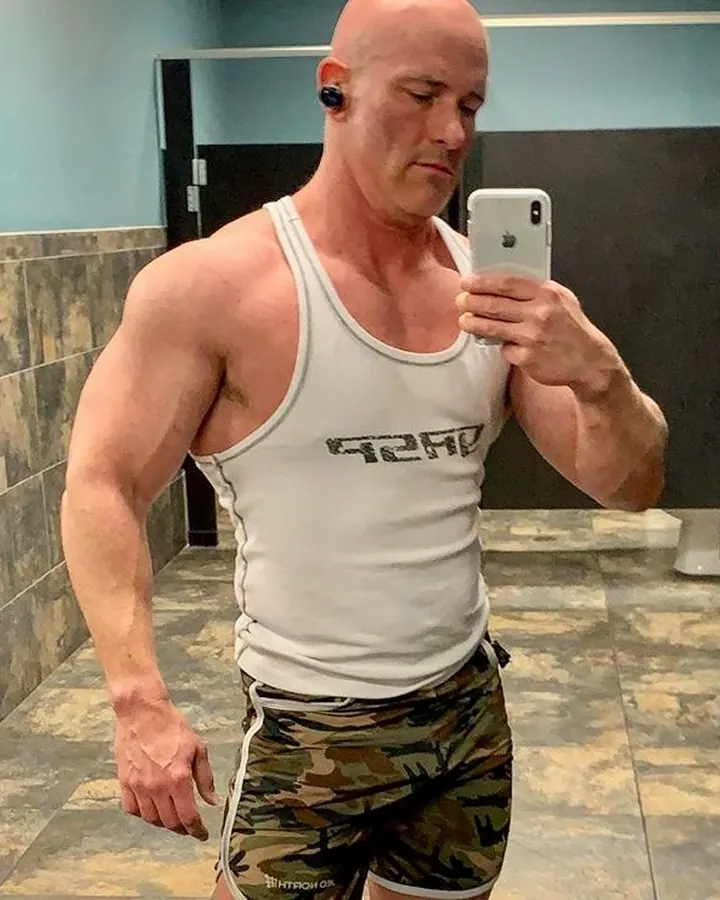 Homme chauve en débardeur blanc, prenant un selfie dans un miroir, regard intense